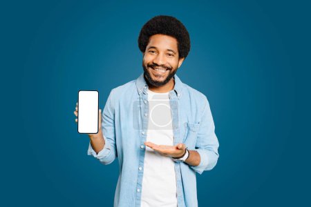 Photo for Handsome young Brazilian man shows an empty smartphone screen with an inviting smile, likely presenting an app or a service. The concept of presentation new app, mock-up - Royalty Free Image
