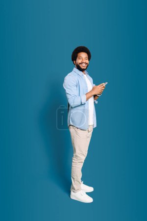 A jovial young man holding a phone and looking at camera, representing the joy of staying connected with friends and family through technology, standing isolated on blue, full length studio shot