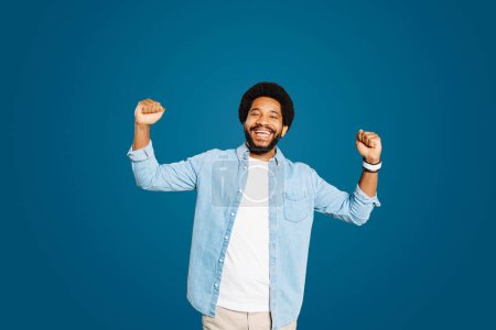 Photo for Young man with cheerful smile raises both fists in a triumphant gesture, showcasing a moment of success or celebration against a blue backdrop. The shoot emphasizes feelings of accomplishment and joy - Royalty Free Image