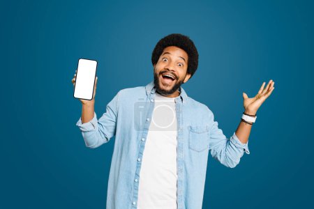 Photo for Happy male freelancer, a young Brazilian man playfully presents his smartphone with a blank empty screen standing isolated on blue, the concept of product reveals or special announcements. - Royalty Free Image