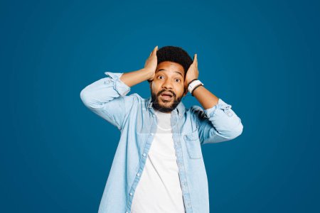 Photo for Young man in a denim shirt looks surprised with hands on his head against blue background. His expression is one of astonishment or having an unexpected idea, adding a sense of drama or humor to image - Royalty Free Image