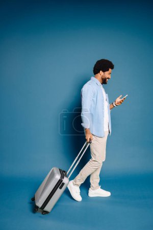 Photo for Cheerful young man with an afro hairstyle is strolling with silver suitcase, attentively looking at his smartphone. The modern traveler navigates through his journey with technology at his fingertips - Royalty Free Image