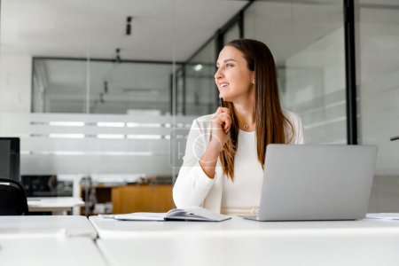 Téléchargez les photos : Une femme d'affaires réfléchit avec le sourire, en regardant loin de l'ordinateur portable, en contemplant une nouvelle idée, ou en réfléchissant à un accord réussi. Concept d'aspects créatifs et stratégiques de la pensée d'entreprise - en image libre de droit