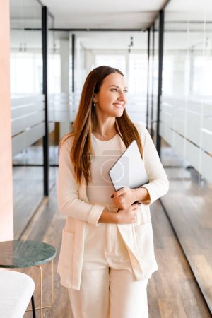 Photo for With a laptop clasped and a contented smile, a young businesswoman walking through the modern office, the serene setting suggesting a well-balanced work life, female office employee in glassy hallway - Royalty Free Image
