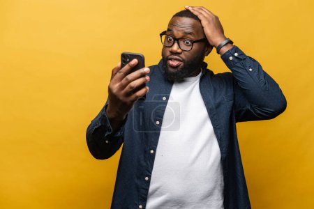 Photo for A surprised African-American man stares at his smartphone with an expression of disbelief, adding drama to the simple action of checking a device, studio shot isolated on yellow - Royalty Free Image