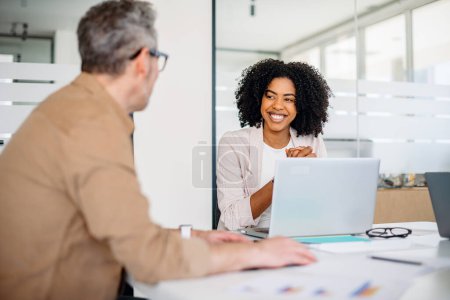 Photo for A joyful female professional with curly hair shares a warm smile while engaging in a productive conversation with her mature male colleague in office, depicting an atmosphere of amicable collaboration - Royalty Free Image
