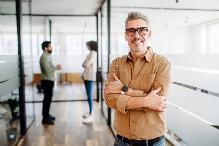 Mature man manager with a genial smile crosses his arms in satisfaction as he stands in his contemporary office space, embodying the spirit of modern professionalism and confident business culture