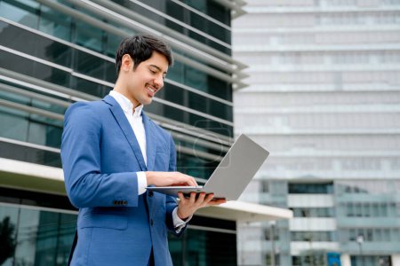 Photo for A smiling Hispanic businessman stands with an open laptop in front of a modern office, embodying the concept of accessible technology and business efficiency in todays fast-paced world. - Royalty Free Image