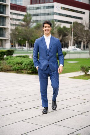 Eine Ganzkörperaufnahme eines fröhlichen hispanischen Geschäftsmannes, der selbstbewusst in einem blauen Anzug geht, wobei eine geschäftige Stadtlandschaft einen dynamischen Hintergrund für seinen aktiven Lebensstil bildet..