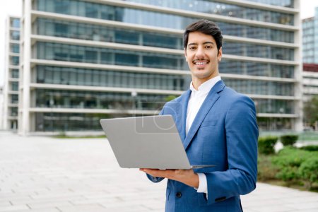 Photo for Confident Hispanic businessman smiles while working on his laptop, symbolizing the intersection of traditional business with modern mobility with a high-rise buildings in the background - Royalty Free Image