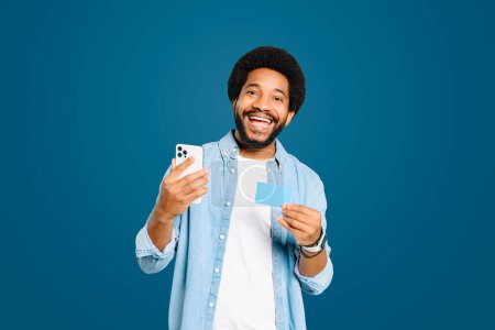 Téléchargez les photos : Homme brésilien excité tient un smartphone dans une main et une carte de crédit dans l'autre, idéal pour les concepts liés à l'achat en ligne ou mobile bancaire debout isolé sur bleu, concept e-banking - en image libre de droit