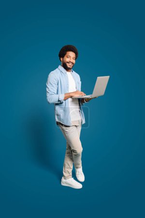 Photo for Smart Brazilian man is full engagement with his laptop on a blue background, full length of a handsome male freelancer working remotely, photo perfect for themes of modern work and connectivity. - Royalty Free Image