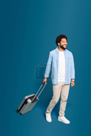 Foto de Una escena dinámica mientras un joven brasileño en traje casual camina con su maleta aislada en azul, estilo de vida activo de un joven viajero en movimiento, listo para nuevas experiencias - Imagen libre de derechos