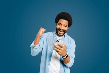 Photo for Happy surprised man laughs and punches the air with his fist, displaying a gesture of success or victory while looking at his phone. The shot embodies the triumph and satisfaction, achieving a goal - Royalty Free Image