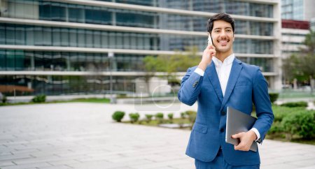 Foto de Un hombre de negocios hispano sonriente con un traje azul hace una llamada en su teléfono inteligente mientras sostiene una computadora portátil, retratando a un empresario exitoso, conectado y móvil en un entorno urbano. Concepto multitarea - Imagen libre de derechos