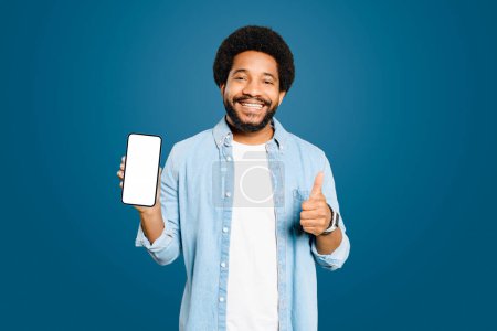 Foto de Un joven brasileño sonriente con camisa de mezclilla que muestra un teléfono inteligente con una pantalla vacía y un pulgar hacia arriba aislado en azul, aprobación y retroalimentación positiva en un contexto digital. - Imagen libre de derechos
