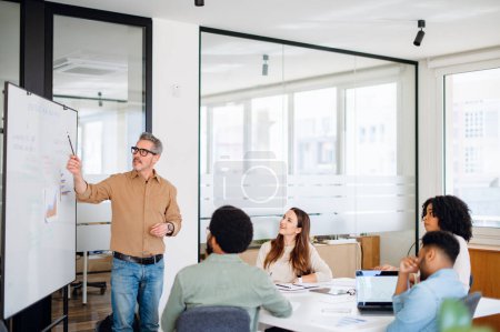 Photo for In a brightly lit office space, mature man with grey hair leads a discussion in front of a whiteboard, highlighting key points with a marker as his diverse team of young professionals listens intently - Royalty Free Image