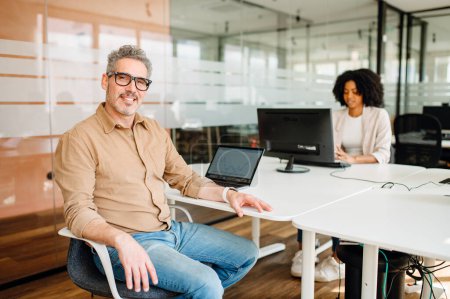 A genial senior business leader sits at an office table with a laptop, engaging in a task with a welcoming posture that suggests openness to collaboration and communication in a modern workplace