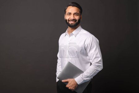 Photo for Indian businessman holds a laptop, presenting a casual yet professional demeanor against a black background, ideal for themes of technology in business - Royalty Free Image