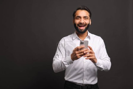 Téléchargez les photos : Un homme d'affaires indien en chemise blanche rayonne de joie en utilisant son smartphone, signe d'une nouvelle positive ou d'une transaction réussie, sur un fond gris foncé - en image libre de droit