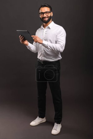 Photo for A cheerful Indian businessman in a white shirt interacts with a digital tablet, his full-body stance and engaging smile conveying accessibility and tech-savviness - Royalty Free Image