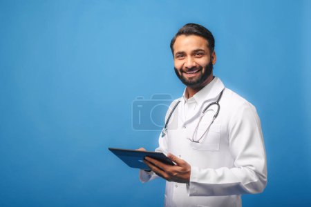 Téléchargez les photos : Un médecin indien au sourire chaleureux porte un blouse blanche avec un stéthoscope autour du cou, tenant en toute confiance une tablette numérique, adaptée aux soins de santé et aux thèmes médicaux. - en image libre de droit
