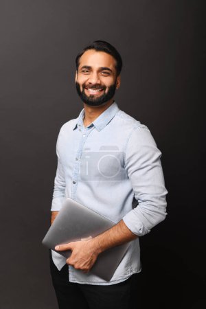 Foto de Un hombre de negocios indio con una camisa azul elegante y casual sostiene un portátil en su brazo, de pie sobre un fondo oscuro con una sonrisa de bienvenida. Retrato de la oficina masculina confiada emlployee - Imagen libre de derechos