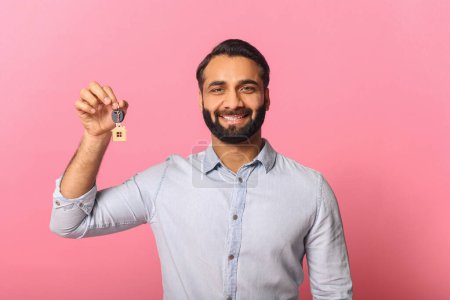 Photo for Against a vibrant pink background, an Indian businessman beams while holding up a house key, symbolizing real estate success and home ownership. The concept of property investment and entrepreneurship - Royalty Free Image