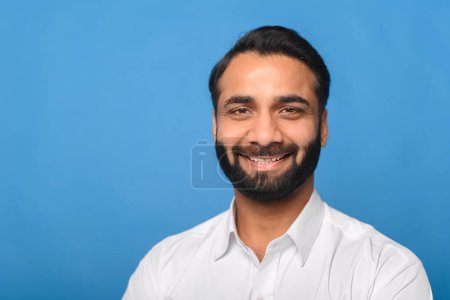 Photo for An Indian man with a friendly smile and a well-groomed beard presents a welcoming expression, wearing a white shirt on a blue backdrop. Concept of customer service, hospitality, or client relations. - Royalty Free Image