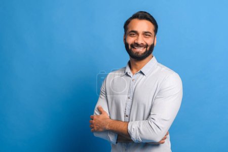 Photo for In front of a bright blue background, the Indian businessman stands with arms crossed, his casual light blue shirt suggesting a relaxed yet confident business approach - Royalty Free Image