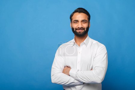 Ein indischer Geschäftsmann in knackigem weißen Hemd steht mit verschränkten Armen vor blauem Hintergrund und strahlt ein professionelles und nahbares Auftreten aus. Geschäfts-, Finanz- oder Unternehmenskonzept