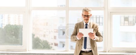 Photo for Mature businessman in a brown suit focuses on a tablet, a portrait of seasoned expertise mingling with modern digital tools, set against a backdrop of bright windows in a spacious office, wide banner - Royalty Free Image