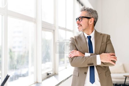 The cheerful mature grey-haired businessman looks off into the distance, arms crossed, standing in a sunny office, confident male manager in stylish eyewear and professional attire