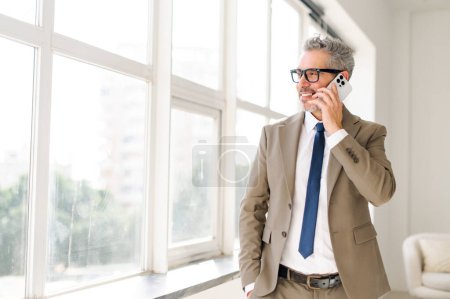 Photo for Senior businessman during a conversation on his phone, looks out window in a stance of thoughtful consideration, standing against an office interior. Concept of importance of connectivity in business - Royalty Free Image
