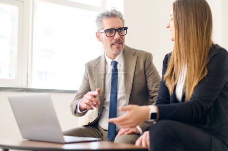 Dans un bureau ensoleillé, un homme d'affaires mature aux cheveux gris écoute attentivement sa collègue pendant qu'elle explique un concept, présentant un échange d'idées et un engagement professionnel..