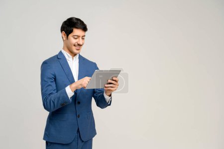 Photo for Young businessman in a business suit interacts with a digital tablet, his expression one of pleasant focus, perfect for illustrating the integration of modern technology in everyday professional tasks - Royalty Free Image