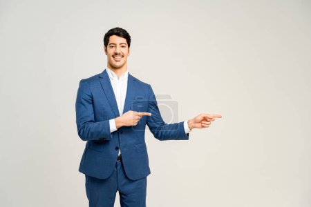 Un homme d'affaires charmant montre le côté avec un sourire confiant, présentant un produit ou un concept d'une manière propre et directe, debout isolé sur le blanc