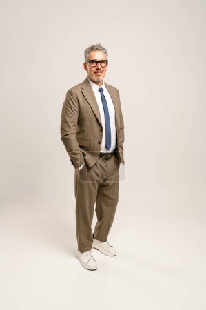 Full body portrait of a senior businessman in a beige suit and casual white sneakers, standing confidently with hands in pockets, blending traditional business attire with a modern twist.