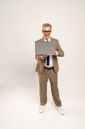 Photo for In a full-length the mature businessman holds a laptop, dressed in a casual yet professional suit and sneakers. This relaxed but professional image perfectly suits themes of modern business agility - Royalty Free Image