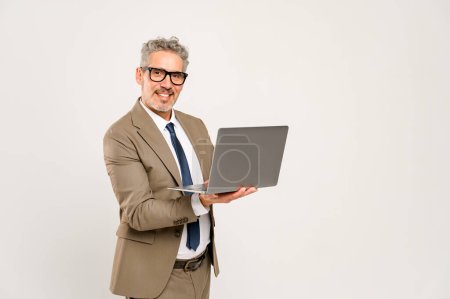 Photo for The grey-haired businessman in a formal suit and glasses standing with the laptop and looking at the camera. This image is ideal for depicting online business management, concentrated work - Royalty Free Image
