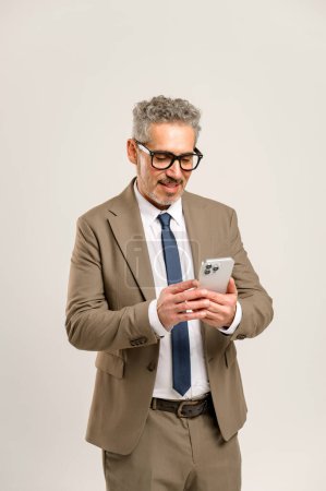 Téléchargez les photos : Un homme d'affaires senior en costume et lunettes utilise un smartphone, représentant l'engagement avec la technologie et la connectivité des cadres modernes dans le monde des affaires. - en image libre de droit