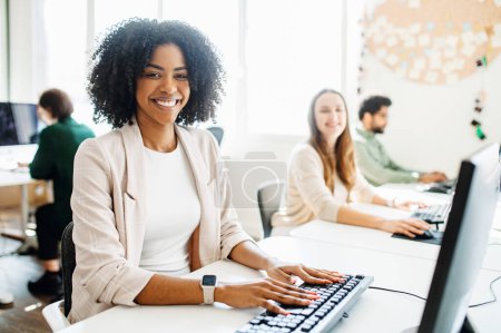 A cheerful female employee with curly hair radiates positivity, as she engages with her work on a desktop computer in an open space office, demonstrating a productive and friendly workplace ambiance