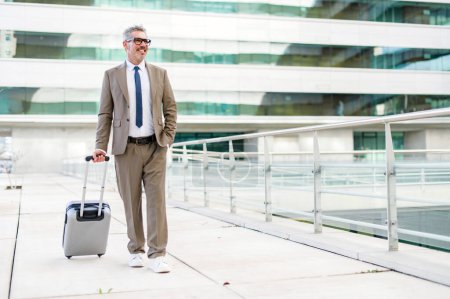 Photo for Mature grey-haired businessman strides confidently, suitcase in tow, against an urban backdrop, representing a professional seamlessly blending travel and business - Royalty Free Image