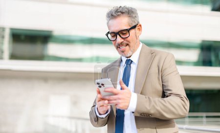 Téléchargez les photos : Homme d'affaires senior intelligent souriant en regardant son smartphone, en examinant une transaction réussie ou un message chaleureux. L'immeuble de bureaux moderne derrière lui fournit une toile de fond professionnelle - en image libre de droit