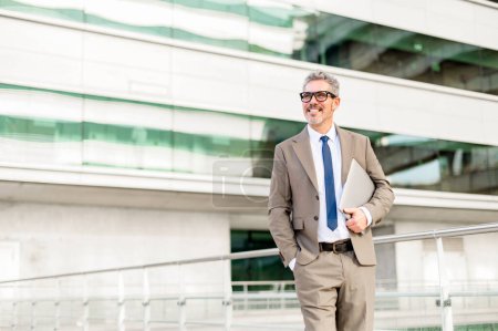 Photo for A content senior businessman holding a laptop walks outdoors, his relaxed demeanor and the modern office backdrop illustrating the flexibility of modern executives to work from anywhere - Royalty Free Image