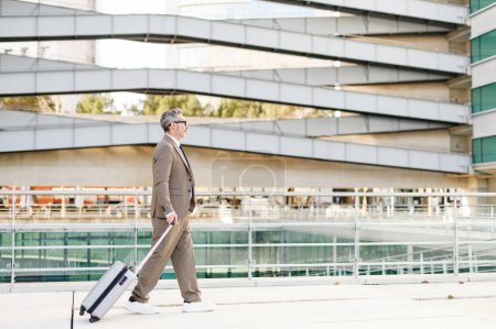 Téléchargez les photos : Un homme d'affaires chevronné marche intentionnellement avec une valise roulante, illustrant la mobilité et la nature rapide de la vie d'entreprise moderne, vue de côté avec bureau en arrière-plan - en image libre de droit