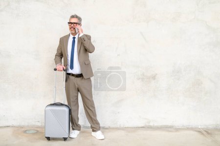 Téléchargez les photos : Un homme d'affaires sénior heureux discute sur son téléphone portable tout en se tenant debout avec sa valise, démontrant la connectivité constante des professionnels mobiles - en image libre de droit
