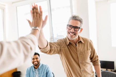 A high-five between colleagues, mature man in focus, emphasizes the exuberant success and positive reinforcement prevalent in their modern workspace. The high energy and team-centric atmosphere