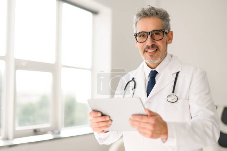 Der leitende Arzt mit Brille und freundlichem Auftreten hält eine Tablette in der Hand und steht an einem Fenster mit natürlichem Licht, das eine moderne und zugängliche Umgebung für das Gesundheitswesen suggeriert. Moderne medizinische Praxis