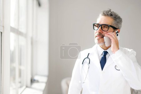 Téléchargez les photos : Un médecin chevronné en manteau blanc est vu s'engager dans une conversation téléphonique par une fenêtre lumineuse. Le concept saisit un professionnel de la santé dans un moment de communication, discuter d'un soin aux patients - en image libre de droit
